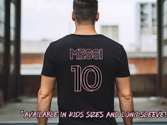 Join the Messi fan club and welcome his new team with this cool t-shirt. A minimalist graphic tee inspired by the uniform of his new team and customized with Messi's details. This tee celebrates Messi's move to Miami and showcases his status as the legend and greatest of all time (GOAT). The shirt is available in adult and kids sizes, and it can be ordered as matching parents and child tee for the ultimate sports apparel.  Designed with a minimalist and unisex style, the tee has a modern vibe, m Jersey T-shirt With Team Name For Fan Merchandise, Football Season Fan Merchandise T-shirt With Logo Print, Custom Logo Crew Neck T-shirt For Sports Events, Sporty Tops With Logo Print For Fan Events, Pre-shrunk Shirt For Sports Fans, Black Tops With Name Print For Fan Gear, Team Name Jersey T-shirt For Fan Merchandise, Jersey T-shirt With Logo Print For Sports Events, Black T-shirt With Letter Print For Fan Events