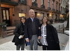 an older man and two women are standing on the sidewalk in front of some buildings