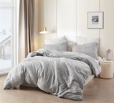 a bed with grey sheets and pillows in a white room next to a large window