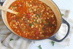 a pot filled with pasta and meat soup on top of a white cloth next to a wooden spoon