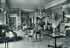 an old black and white photo of people in a living room with chairs, couches, tables and pictures on the wall