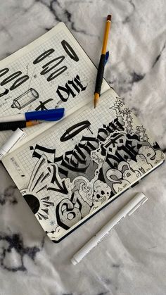 an open notebook with graffiti written on it and two pens next to it, sitting on a marble surface