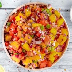 a bowl filled with chopped vegetables next to some lemons and lime wedges on a white wooden table