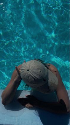 a woman in a hat sitting on the edge of a swimming pool with her hands behind her head