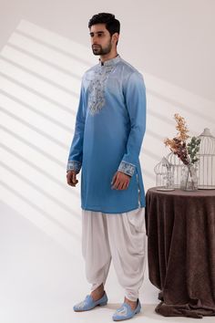 Blue sleeveless front buttons bundi with paisley bloom embroidery using beads and pearls. Paired with long sleeves ombre kurta with floral embroidery on the placket and white cowl pant.
Components: 3
Pattern: Embroidered
Type Of Work: Paisley Bloom
Neckline: Mandarin Collar
Sleeve Type: Bundi: Sleeveless, Kurta: Long
Fabric: Raw Silk, Handloom Silk,  Modal Satin
Color: Blue, White
Other Details: 
Approx weight: 2kgs
Poly lining
Occasion: Mehendi and Puja, Sangeet - Aza Fashions Blue Cotton Nehru Jacket For Wedding, Fitted Blue Nehru Jacket With Floral Embroidery, Blue Nehru Jacket With Resham Embroidery, Ceremonial Blue Embroidered Nehru Jacket, Blue Embroidered Cotton Nehru Jacket, Designer Long-sleeve Embroidered Nehru Jacket, Bloom Embroidery, Sleeveless Kurta, Kurta Set For Men