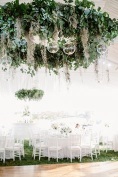 an indoor wedding with white chairs and greenery hanging from the ceiling, surrounded by round chandeliers