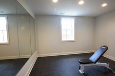 an empty gym room with a bench and mirror in the corner, along with two windows