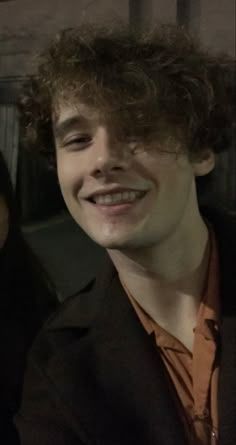 a young man with curly hair smiling at the camera while wearing an orange shirt and black blazer