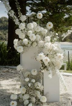 an arrangement of white flowers in a vase on the ground near some trees and water