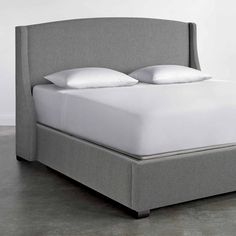 an upholstered bed with two pillows and white sheets on it, in front of a gray wall