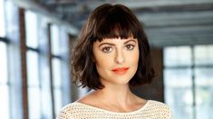 a woman with dark hair and orange lipstick looks at the camera while wearing a white sweater