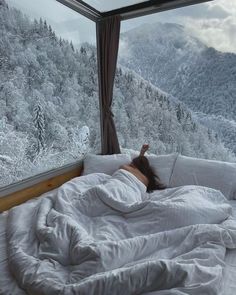 a woman is laying in bed looking out the window at snow covered mountains and trees