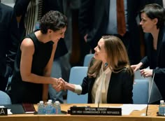 two women shaking hands in front of other people at a conference room table with microphones