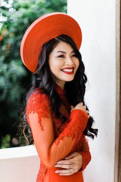 a woman in an orange dress and red hat smiles at the camera while leaning against a wall