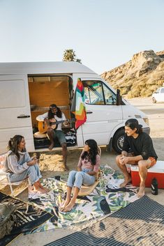 four people sitting on the beach in front of a van