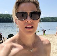 a woman wearing sunglasses on the beach near some water and people in the background looking at the camera