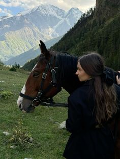 a woman standing next to a brown horse on top of a lush green hillside covered in snow capped mountains