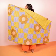 a woman holding up a large yellow and purple flowered blanket over her head while standing in front of a pink wall