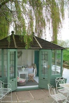 an outdoor room with green doors and white chairs on the patio next to a pond