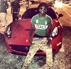 a man sitting on the hood of a red sports car in front of some people