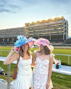 two women standing next to each other in front of a race track wearing matching hats