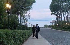 a man and woman walking down a path next to the ocean at dusk with lights on