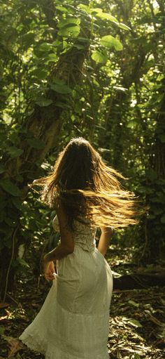 a woman in a white dress is walking through the woods with her hair blowing in the wind