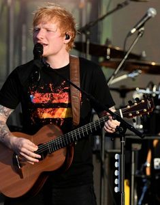 a young man with red hair playing an acoustic guitar and singing into a microphone on stage