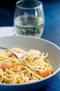 a white bowl filled with pasta and shrimp on top of a blue table next to a glass of wine