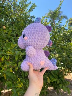 a hand holding a purple crocheted teddy bear in front of some bushes and trees