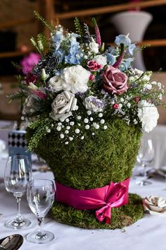 an arrangement of flowers and greenery in a moss covered basket on a table with wine glasses