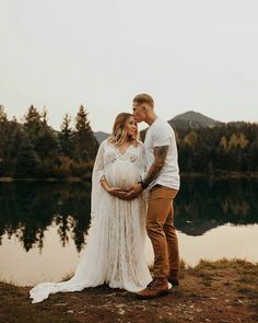 a pregnant couple standing next to each other in front of a lake