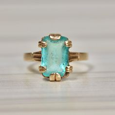 10K Yellow Gold, Vintage Retro Turquoise Ring, Synthetic Stone in bold setting, Size 8 Weight:  5.00 grams Ring Size:  8 Stone Measurements:  11.80 mm x 7.75 mm Shank:  2.55 mm This striking vintage ring, dating back to the 1930s or 1940s, features a captivating synthetic emerald cut stone set in a bold, retro-style setting. Its bright turquoise hue, reminiscent of old Hollywood glamour, is beautifully offset by the ring's chunky, statement-making design. Though the stone bears a small chip in the bottom right corner and light abrasion from age, these imperfections only add to its unique character, telling the story of its past. An estate piece with timeless charm, this ring effortlessly captures the essence of vintage elegance while making a daring, unforgettable statement. Vintage Yellow Gold Turquoise Ring For Formal Occasions, Vintage Turquoise Ring In 14k Gold, Vintage Yellow Gold Turquoise Ring For Anniversary, Turquoise Emerald-cut Gemstone Ring, Turquoise Gemstone Rings With Emerald Cut, Turquoise Emerald Cut Gemstone Ring, Emerald Cut Turquoise Gemstone Ring, Vintage Oval Aquamarine Ring, Vintage Aquamarine Oval Ring