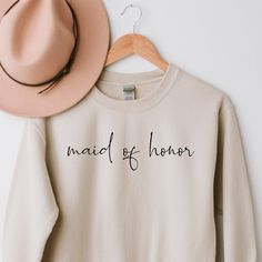 a white sweatshirt with the words maid of honor printed on it next to a pink hat