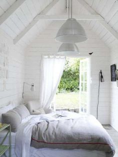 a bed sitting under a window in a bedroom next to a table and chair with a lamp hanging from the ceiling