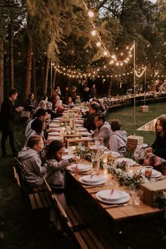 a group of people sitting around a long table with plates and glasses on it, surrounded by string lights