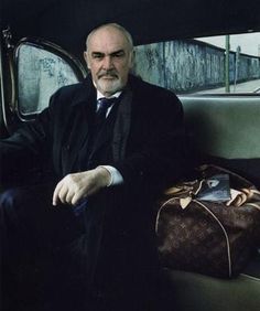 a man sitting in the back seat of a car next to a brown bag and purse