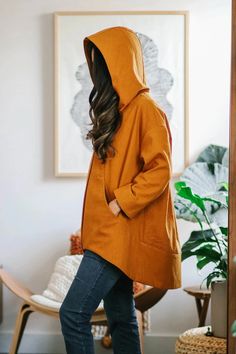 a woman in an orange hoodie is standing near a chair and potted plant