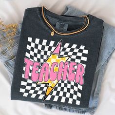 Get ready to rock the classroom with our Checkered Lighting Bolt Pencil Teacher Shirt, a retro-inspired design perfect for your first day of school! Embrace the back-to-school vibes in comfort with our stylish Comfort Colors Shirt. 📚 Shine Bright on Day One! 🌟 Celebrate the first day with our Checkered Lighting Bolt Pencil Teacher Shirt! This retro-inspired Back to School tee is not just a shirt--it's your statement piece for the classroom. Made with cozy Comfort Colors, it's perfect for all-d Retro Cotton Shirt With Letter Print, Retro Letter Print T-shirt For School, Retro Graphic Print Shirt For School, Retro Black T-shirt For School, Retro Graphic Print Tops For School, Retro Style Tops For End Of School Year College, Retro Black Top For School, Trendy Graphic Print Tops For School, School Spirit Cotton Shirt With Graphic Print