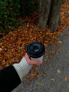 a person holding a coffee cup in their hand on a sidewalk with leaves all over the ground