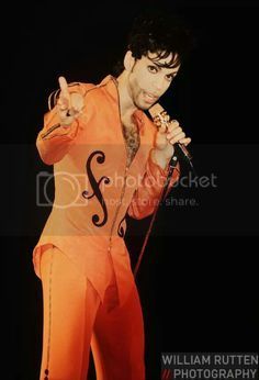 elvis presley performing on stage in an orange suit with his hands up and holding a microphone