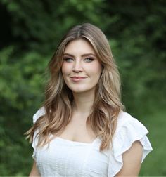 a beautiful young woman standing in front of green grass and trees wearing a white top