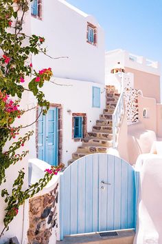 a white building with blue doors and steps leading up to the door is surrounded by pink flowers