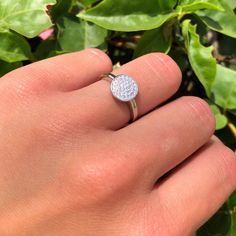 a person's hand holding a ring with a diamond in the middle and green leaves behind it