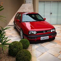 a red car parked in front of a house next to green plants and bushes on the sidewalk