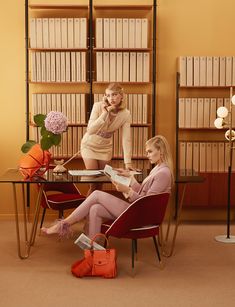 two women are sitting at a table in front of bookshelves, one is talking on the phone