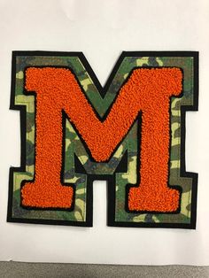 the letter m is made out of orange and camo material with black trim on it