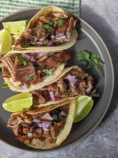 three shredded pork tacos on a plate with limes and cilantro wedges