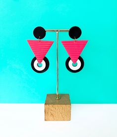 two pink and black triangle shaped earrings sitting on top of a wooden stand against a blue wall