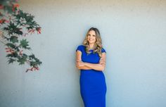 a woman in a blue dress leaning against a wall with her arms crossed and smiling